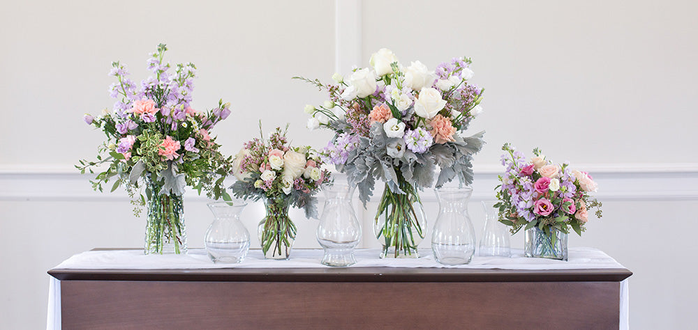  A table with several different vases and pastel colored floral arrangements.