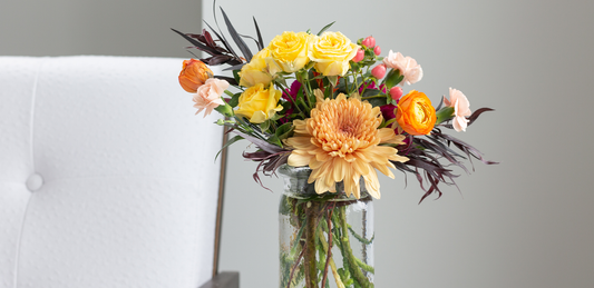 A bouquet of orange and yellow flowers in a clear jar vase on a side table